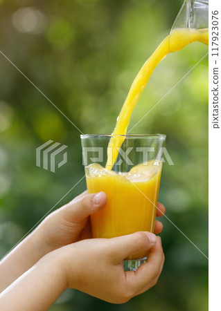 child hands holding glass and orange juice pouring from jug with green garden on the background 117923076