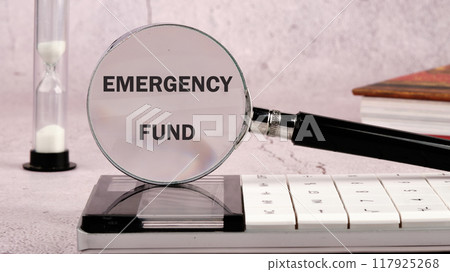 Concept of setting money saving goal for rainy day. EMERGENCY FUND through a magnifying glass on a gray background in a composition with a calculator, an hourglass and a book 117925268
