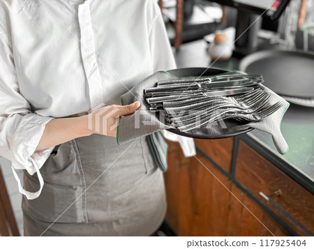 Waiter in white shirt holding black tray with neatly arranged silverware including forks and knives on grey napkin, ready for table setting, with blurred background of restaurant interior. 117925404