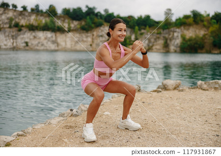 A Young Woman Squatting Gracefully by a Scenic Lake While Dressed in Trendy Athletic Wear 117931867