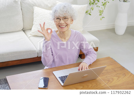 Senior woman making an OK sign in front of a laptop computer in living room Senior woman making an OK sign in front of a laptop computer in living room 117932262