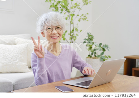 Senior woman making a peace sign in front of a laptop in the living room Senior woman making a peace sign in front of a laptop in the living room 117932264