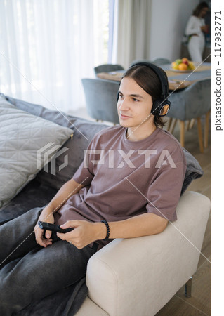 Teenage boy watch tv in wireless headphones. Male siting in living room. Background of sister in kitchen Teenage boy watch tv in wireless headphones. Male siting in living room. Background of sister in kitchen 117932771