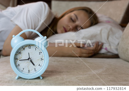 Alarm clock on sofa will soon wake up the young caucasian girl in the background who is lying on pillow during nap, control of human sleep 117932814