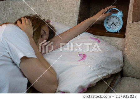 Young woman reaches to turn off alarm clock on shelf after nap covering her ear from annoying alarm sound, hard awakening after deep sleep at home 117932822