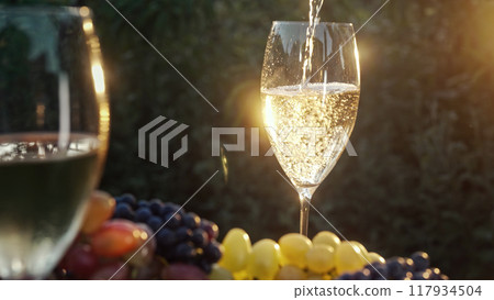 Stunning view of pouring white wine into glass goblet with fresh grape bunch background, sun light 117934504