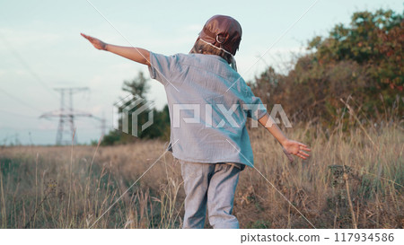 Happy boy in aviator pilot helmet runs as airplane on nature meadow, outside city. Kids dream is to 117934586