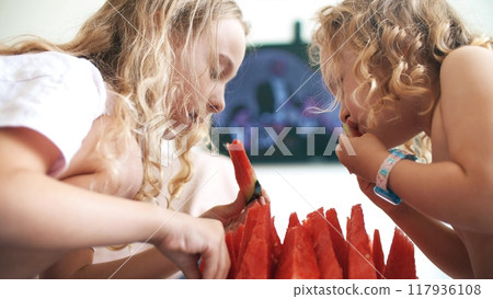 Little sisters eat juicy watermelon slices and watch TV at home 117936108