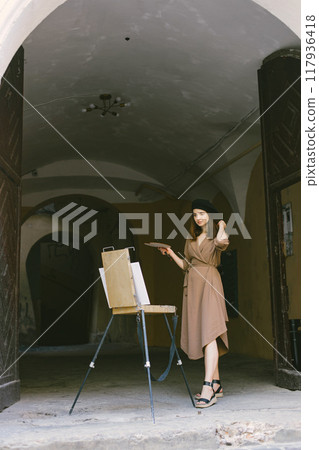 Female painter painting in the city. A woman wearing khaki dress and a black hat. Girl drawing a picture on an easel in summer on a street. Female painter painting in the city. A woman wearing khaki dress and a black hat. Girl drawing a picture on an easel in summer on a street. 117936418