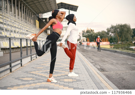 Two women on a stadium doing stretching. Muslim woman in hijab wearing white shirt and red trousers. Brunette woman wearing rose top and black leggins. Two women on a stadium doing stretching. Muslim woman in hijab wearing white shirt and red trousers. Brunette woman wearing rose top and black leggins. 117936466