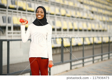 Girl working out outdoors on a stadium. Muslim woman wearing white shirt and red trousers. Girl doing excercises with a dumbbells. Girl working out outdoors on a stadium. Muslim woman wearing white shirt and red trousers. Girl doing excercises with a dumbbells. 117936473