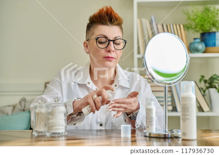 Middle aged woman looking in mirror, using face creams, cosmetic care products 117936730