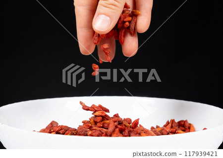 Hand pouring dried goji berries into a plate on a black background. Hand pouring dried goji berries into a plate on a black background. 117939421