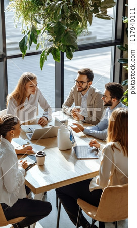 Diverse team collaborating around a table in a bright office with greenery. Business meeting, teamwork, and corporate planning. 117940111