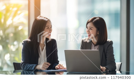 Two Asian businesswomen discussing work in a bright and modern office environment. Professional teamwork and corporate strategy. 117940300