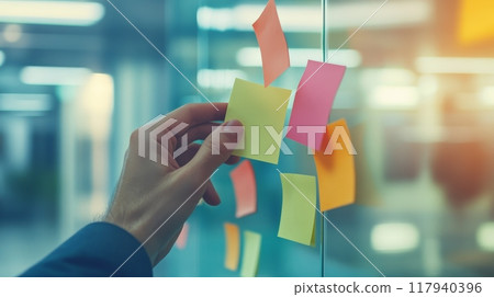 Close-up of a hand placing a pink sticky note on a glass wall in a modern office, symbolizing creativity and brainstorming. 117940396