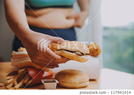 To eat or not. Cheerless plump woman looking at the burger while thinking to eat it or not 117941014