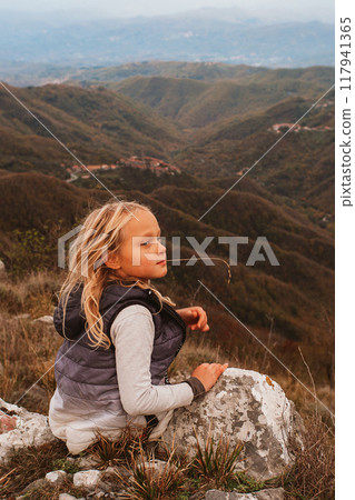 a little blonde girl resting on a stone during a hike in the mountains against the backdrop of an autumn mountain landscape a little blonde girl resting on a stone during a hike in the mountains against the backdrop of an autumn mountain landscape 117941365