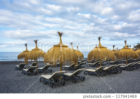 Empty sun beds and straw umbrellas on a beach of pebbles. 117941469