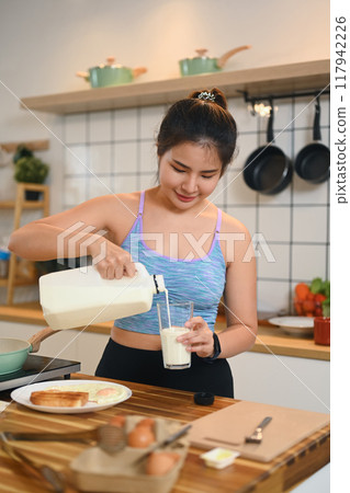 Attractive young woman pouring milk into a glass while standing in a kitchen 117942226