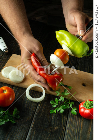 Preparing a vegetable mix on the kitchen table for dinner. The hands of the chef thread ripe red bell peppers onto a fork for grilling 117943217