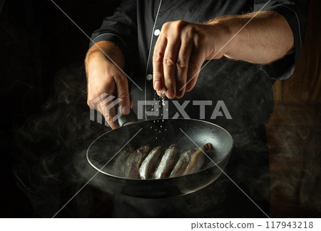 Adding salt to fish in a frying pan. Chef cooking capelin in the hotel kitchen. Concept on a dark background of preparing fish menu 117943218
