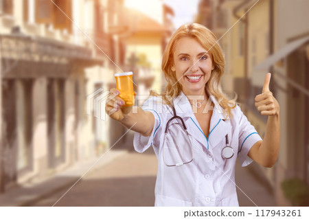 Portrait of happy caucasian female doctor standing outdoors holding a can of pills and gives her thumb up. European city street in the background. Portrait of happy caucasian female doctor standing outdoors holding a can of pills and gives her thumb up. European city street in the background. 117943261