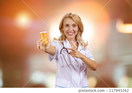 Portrait of happy blonde caucasian mature female doctor pointing at orange bottle of pills. Blurred glowing lights in the background. 117943262