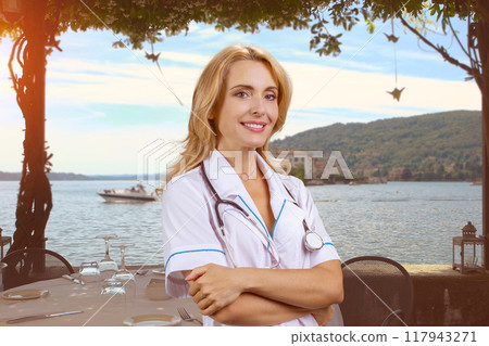 Portrait of happy cheerful female blonde doctor with stethoscope standing outdoors with folded arms. Beach in the background. 117943271