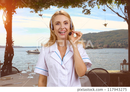 Portrait of blonde mature female doctor wearing headset on the sea coast. Sea beach landscape in the background. Portrait of blonde mature female doctor wearing headset on the sea coast. Sea beach landscape in the background. 117943275