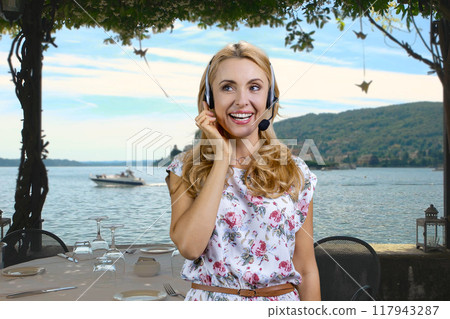 Portrait of mature blonde woman with headset standing outdoors. Sea beach landscape in the background. Portrait of mature blonde woman with headset standing outdoors. Sea beach landscape in the background. 117943287