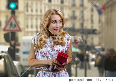 Portrait of happy mature surprised blonde woman taking money from her red wallet. City street in the background. 117943294