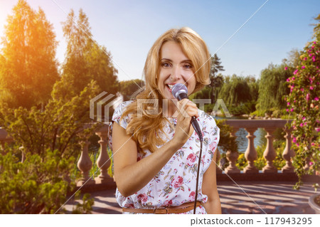 Portrait of blonde woman singing into microphone outdoors. Park trees in the background. 117943295