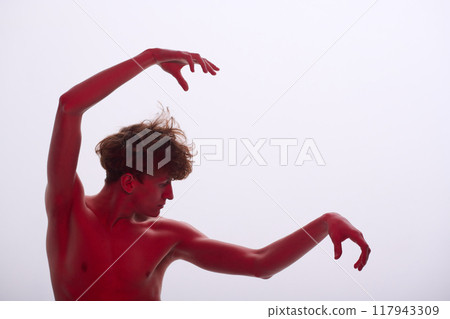 Individual performing elegant dance movements with raised arms, creating dynamic shapes against minimalistic backdrop. Red lighting adds dramatic effect to the performer's pose 117943309