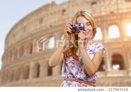Mature blonde woman taking a photo using retro vintage photo camera. Ancient coliseum in the background. Mature blonde woman taking a photo using retro vintage photo camera. Ancient coliseum in the background. 117943310