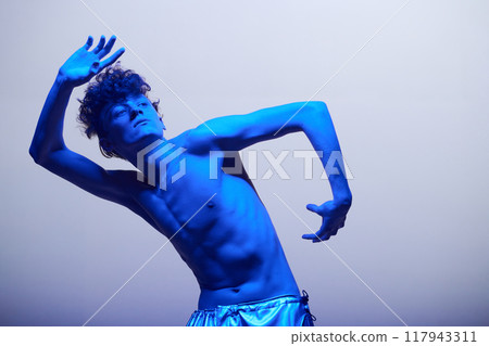 Shirtless man dancing and posing under bright blue lighting creating dramatic effect. Artistic, expressive performance partially obscured by shadows in minimalist setup Shirtless man dancing and posing under bright blue lighting creating dramatic effect. Artistic, expressive performance partially obscured by shadows in minimalist setup 117943311