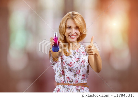 Portrait of happy blonde mature woman holding a perfume bottle and giving her thumb up. Abstract blurred background. 117943315