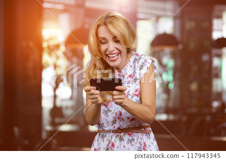 Happy blonde mature woman playing online video game on her smartphone indoors. Home interior in the background. 117943345