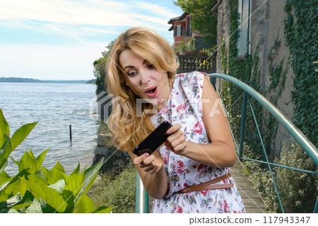 Mature blonde woman playing a video game on her smartphone on vacation. Lake landscape in the background. 117943347