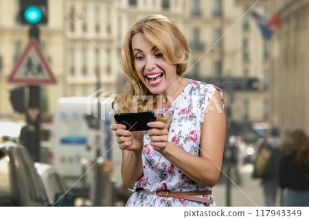 Portrait of mature blonde woman plays video game outdoors. City street in the background. 117943349