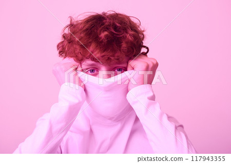 Individual with curly hair hiding behind shirt, gazing intently with visible eyes. Pinkish lighting adding unique flair to the image Individual with curly hair hiding behind shirt, gazing intently with visible eyes. Pinkish lighting adding unique flair to the image 117943355
