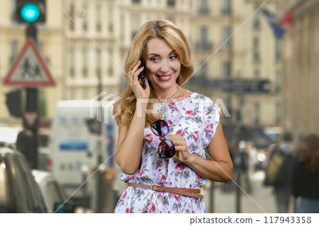 Portrait of excited mature woman wearing with sunglasses talking on phone outdoors. Urban city street in the background. Portrait of excited mature woman wearing with sunglasses talking on phone outdoors. Urban city street in the background. 117943358