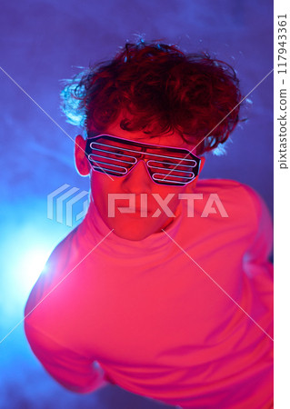 Portrait of person looking straight at camera, wearing futuristic LED glasses and a white turtleneck, against a vibrant background emitting blue and pink lights Portrait of person looking straight at camera, wearing futuristic LED glasses and a white turtleneck, against a vibrant background emitting blue and pink lights 117943361