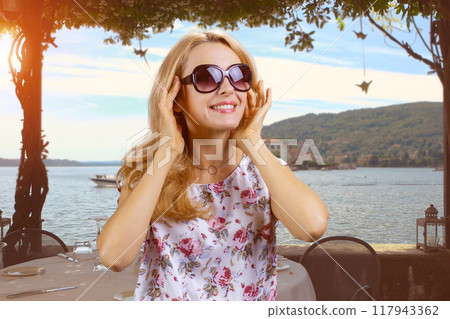 Happy cheerful mature woman wearing sunglasses on vacation. Evening beach seascape in the background. 117943362