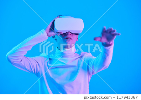 Person immersed in virtual reality experience, wearing VR headset and reaching out with hand against blue background. Portrait of person engaging with futuristic technology 117943367