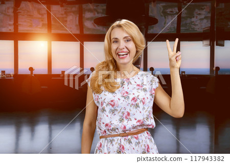 Portrait of charming blonde mature woman shows victory gesture sign indoors. Evening sunshine from the outdoors. Portrait of charming blonde mature woman shows victory gesture sign indoors. Evening sunshine from the outdoors. 117943382