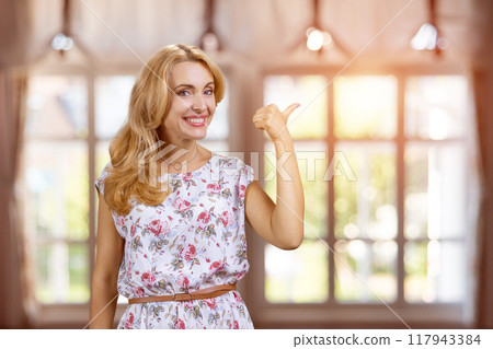 Portrait of attractive blonde mature woman pointing back with her thumb. Indoor window in the background. Portrait of attractive blonde mature woman pointing back with her thumb. Indoor window in the background. 117943384
