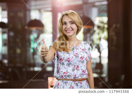 Portrait of charming mature blonde lady shows her thumb up. Restaurant indoors in the background. Portrait of charming mature blonde lady shows her thumb up. Restaurant indoors in the background. 117943388