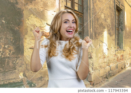 Portrait of excited mature woman standing outdoors celebrating something. Shabby facade in the background. 117943394