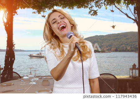 Portrait of happy mature blond woman singing in microphone. Evening beach in the background. 117943436